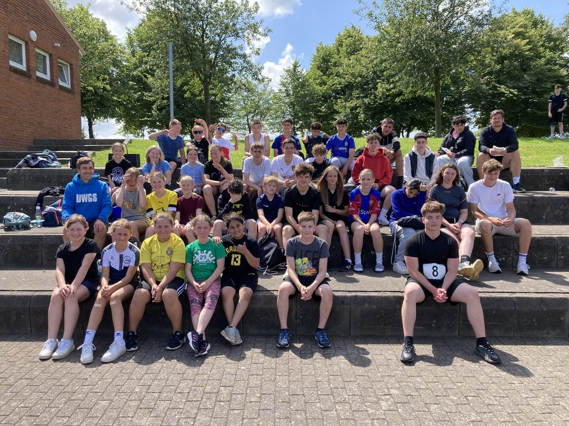 Ca. 50 Schülerinnen und Schüler sitzen bei Sonnenschein auf der Tribüne des Aleestadions in Schleswig.