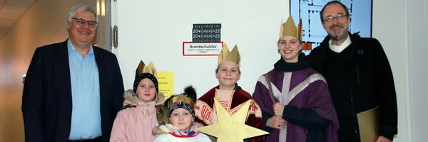 Das Bild zeigt die Sternsinger mit Pastor Winter und dem Bürgermeister der Stadt Schleswig Stephan Dose. Im Hintergrund ist das an eine Tür geklebte Kreidezeichen "20*C+M+B+24" zu erkennen, mit dem der Segen Gottes in das Schleswiger Rathaus gebracht wird.