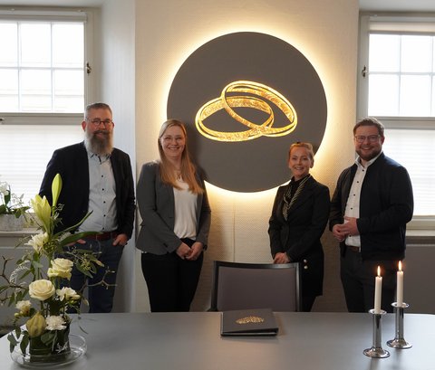 Robert Winterscheidt (Leiter des Standesamts), Kerstin Legant (Standesbeamtin), Fachdienstleiterin Karen Suhling und Bürgermeister Jonas Kähler (v.l.)vor dem beleuchteten Kunstobjekt im Trauzimmer, das zwei goldene Ringe zeigt.
