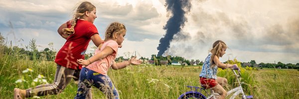 Das Bild zeigt drei Kinder. Rechts fährt ein Mädchen auf einem Fahrrad auf einer Wiese und links laufen zwei Mädchen hinterher. Im Hintergrund ist schwarzer am Horizont aufsteigender Rauch zu sehen. Es ist das UNICEF-Foto des Jahres.