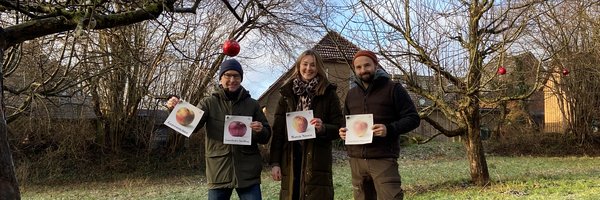 Jens Bagehorn-Delor, (Stadt Schleswig) Maren Thiesen (Naturpark Schlei) und Miklas Staiger (Obstbaumpfleger und Streuobstpädagoge) stehen vor einem Obstbau und halten Schilder mit Äpfeln hoch