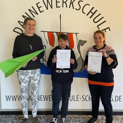 Foto (von links) Schulleiterin A. Schönberg mit Schüler und Lehrkraft D. von Klein vor dem Logo der Dannewerkschule-Schleswig